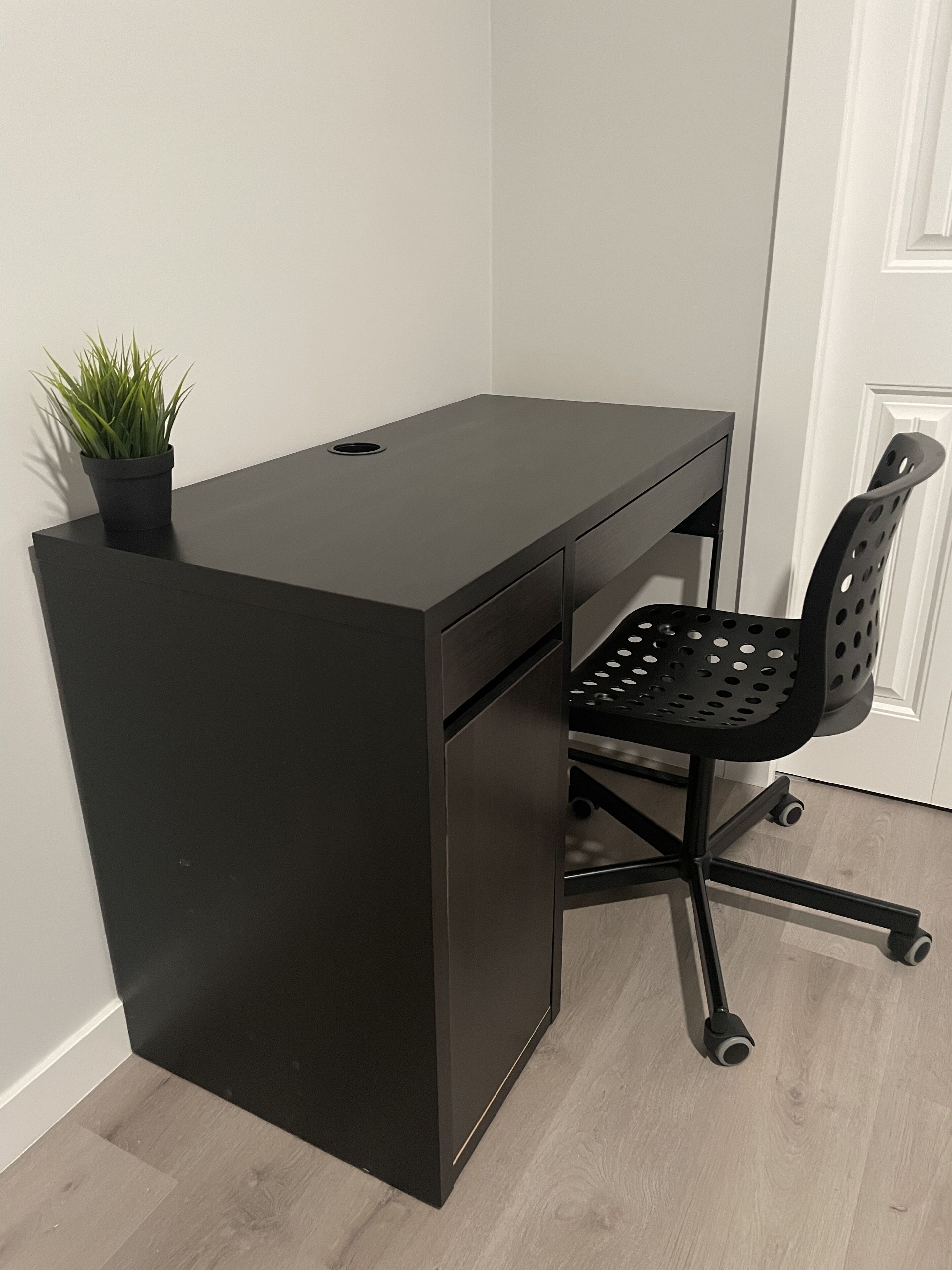 A black desk with a potted plant, a black office chair with wheels, and a white door in the background.