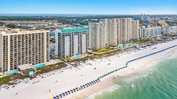Un hotel frente a la playa con varios edificios altos, una playa de arena y una hilera larga de sombrillas.