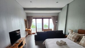A modern hotel room with a large bed, a TV on a wooden stand, a sofa, and a balcony with curtains. - Nelayan Sanur Bay (Bali)