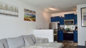 Un salon moderne avec un canapé d’angle gris, une cuisine aux armoires bleues et aux appareils en acier inoxydable, et deux tableaux accrochés aux murs.