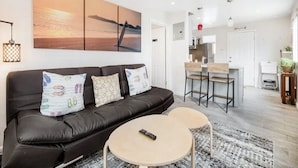A modern living room with a black leather sofa, two round coffee tables, and a large painting above the sofa. - Cozy 1BR Bungalow by the beach, coffee, and dining (Oceanside)