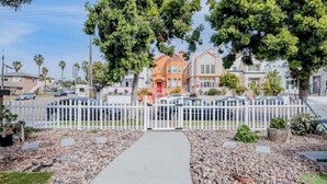 A residential street with a white picket fence, a paved walkway, and houses with visible front yards and trees. - Cozy 1BR Bungalow by the beach, coffee, and dining (Oceanside)