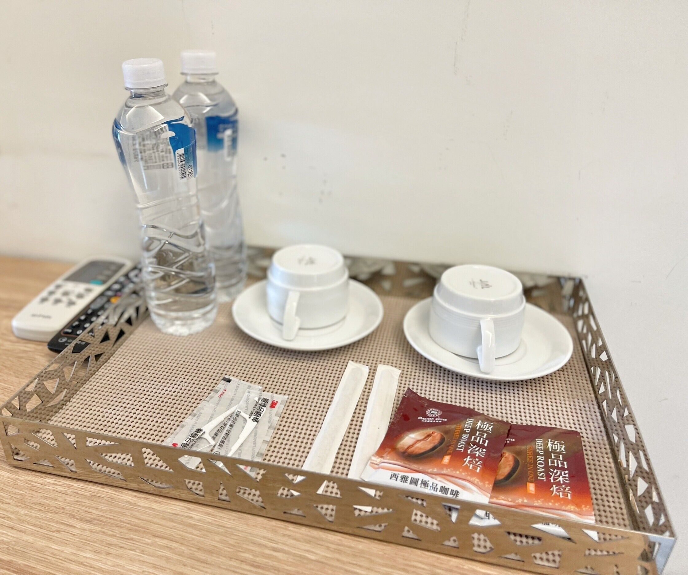 Una bandeja con un control remoto, dos botellas de agua, dos tazas blancas y un sobre de leche para café sobre un tapete tejido.