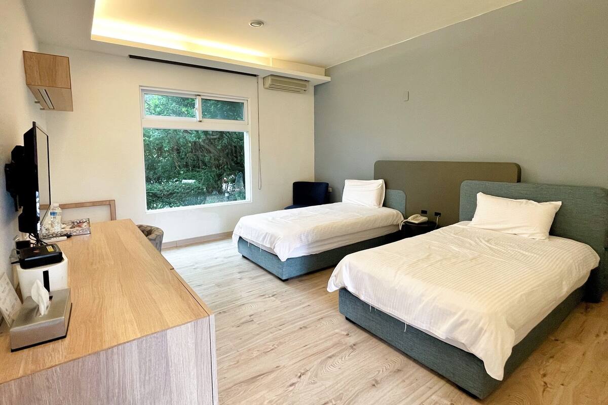 Una camera d'hotel moderna con due letti, una scrivania e un'ampia finestra con vista sugli alberi.