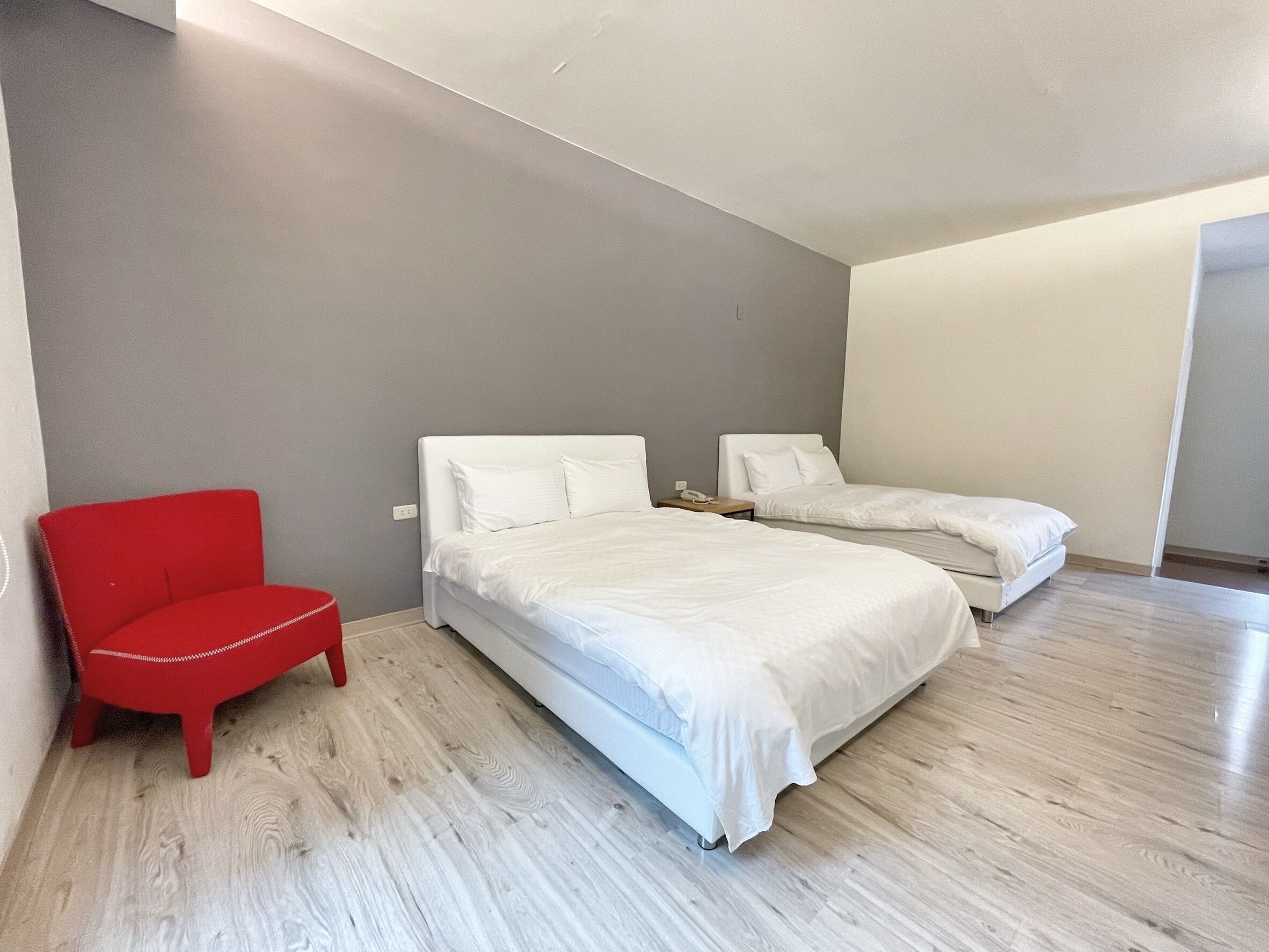 A modern bedroom with a large bed, a smaller bed, a red chair, and a bedside table with a phone.