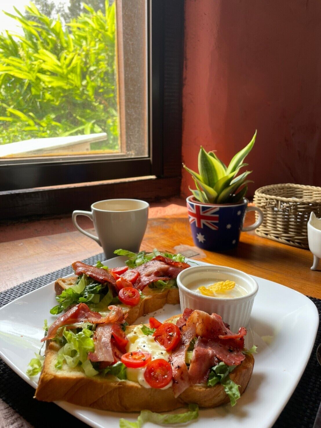 Un plato de desayuno con tostadas cubiertas de tocino, tomates y verduras de hoja, una taza pequeña de sopa y una taza con motivos de bandera.