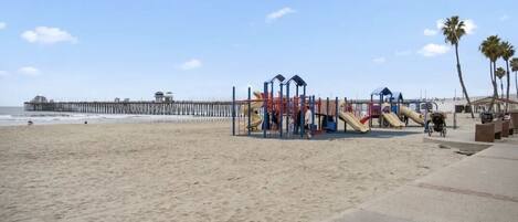 A beach with a playground, a pier in the background, and palm trees.