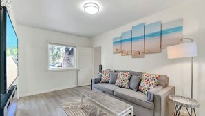 Een moderne woonkamer met een grijze bank, gestreepte kussens, een salontafel en een vloerlamp. Drie schilderijen aan de muur beelden strandtaferelen af.