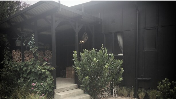 Un edificio con un portico coperto, un muro di pietra con tronchi di legno e un piccolo albero in primo piano.