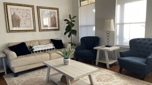 Een woonkamer met een beige bank, twee fauteuils, een salontafel en een grote plant. Twee kunstwerken hangen in lijsten aan de muur en er is een raam met zonwering.