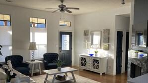 Een ruime woonkamer met een donkerblauwe fauteuil, een witte salontafel en een witte bank. Er is een plafondventilator, een raam met zonwering en een deur met ruitverdeling.