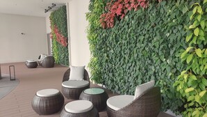 Un área de estar al aire libre moderna con sillas de mimbre y mesas redondas, rodeada de paredes de follaje verde y rojo.