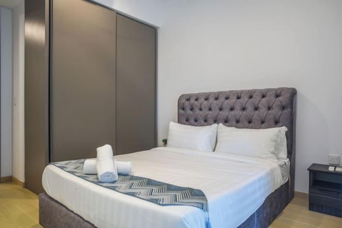 A neatly made bed with a patterned bedspread and pillows, a dark-colored wardrobe, and a small bedside table with a lamp.