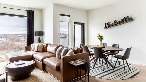 A modern living room with a leather sofa, a dining table with chairs, and a large window offering a city view.