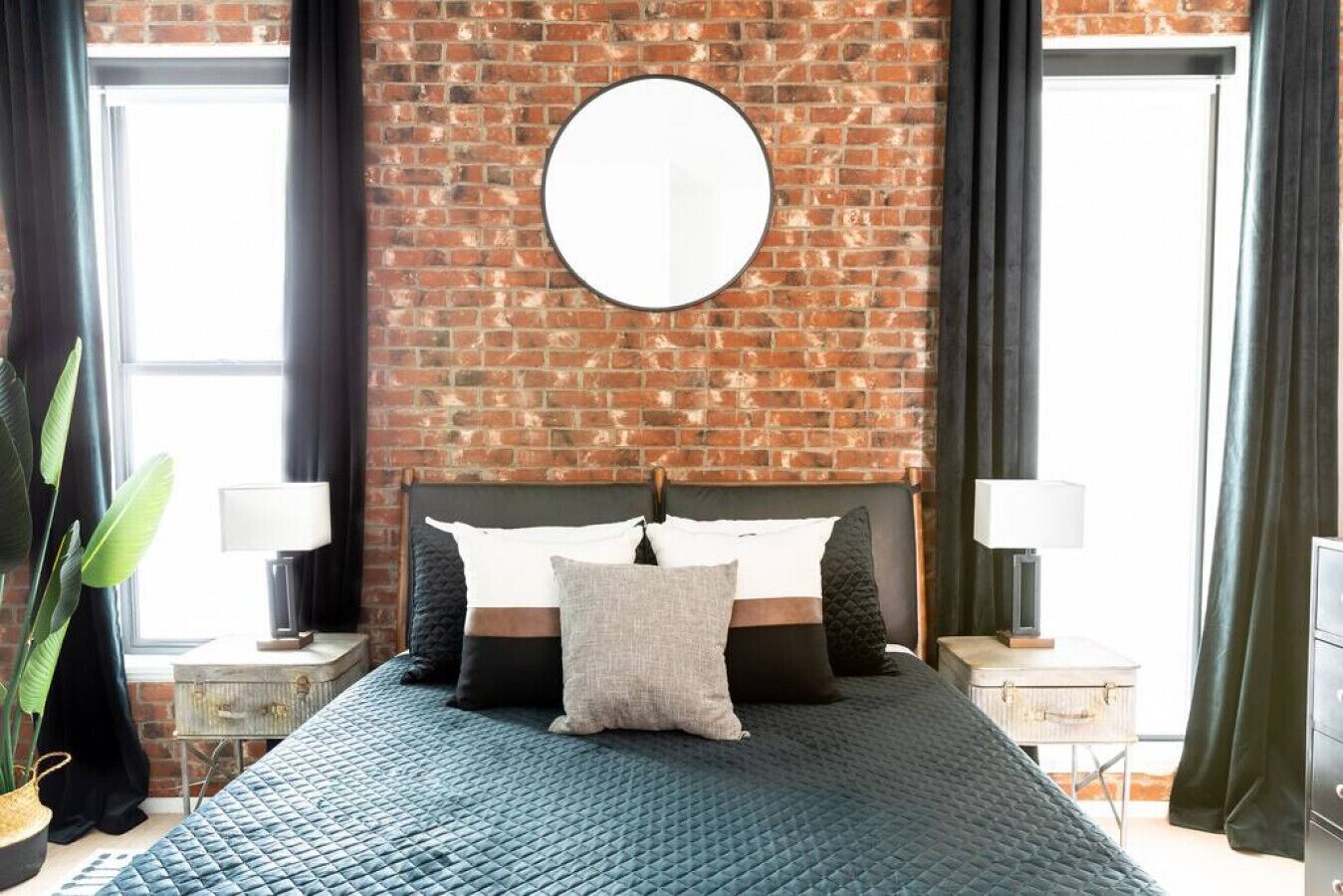 Un dormitorio con pared de ladrillo, un espejo redondo, una cama con varias almohadas, dos mesitas de noche con lámparas y un ventanal con cortinas.