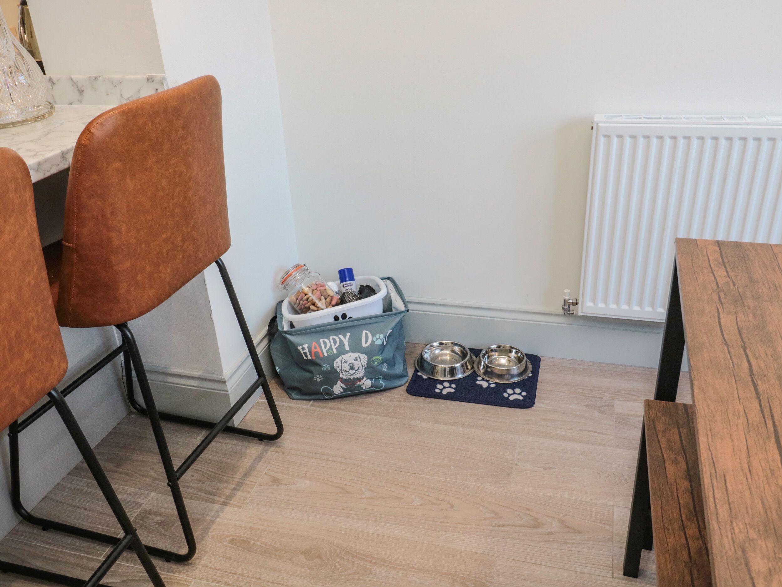 Een eetgedeelte met een houten tafel, twee bruine stoelen, een zak voor diervoeding, twee metalen voerbakken en een marmeren aanrecht.