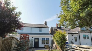 A white two-story house with a black door, a small garden with a wooden table and chairs, and a wooden fence. - 9 Station Cottages (Belford)