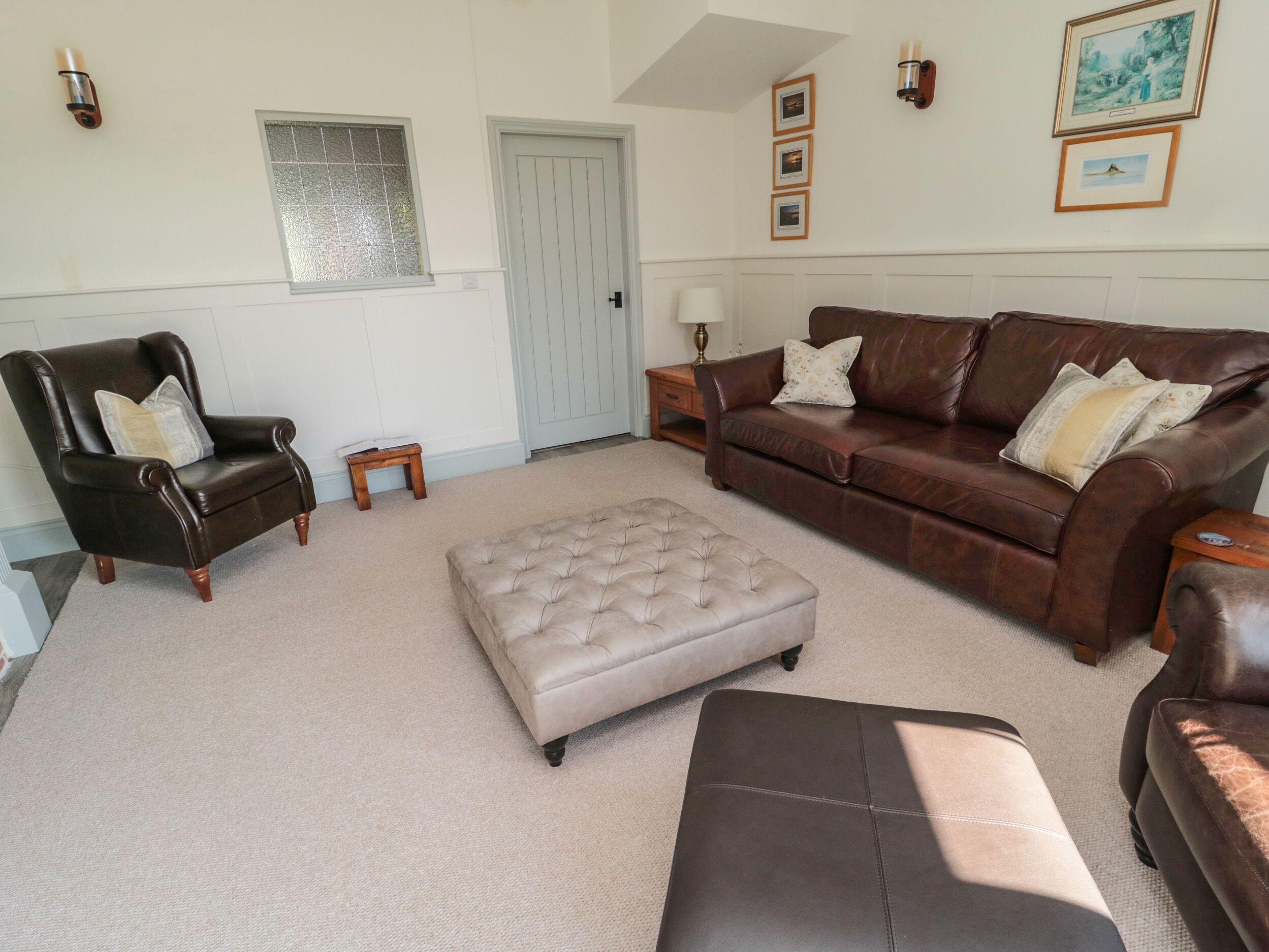 A living room with a brown leather armchair, a brown leather sofa, and a beige ottoman. There is a small wooden table and a lamp. The room has white walls, a door, and a window with a textured glass panel.