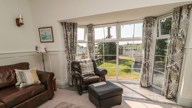 A living room with a leather sofa, a tufted ottoman, and a leather armchair with a cushion. A large window with patterned curtains offers a view of a garden. - 9 Station Cottages (Belford)