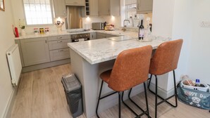A modern kitchen with a central island, two bar stools, and a small trash bin. - 9 Station Cottages (Belford)
