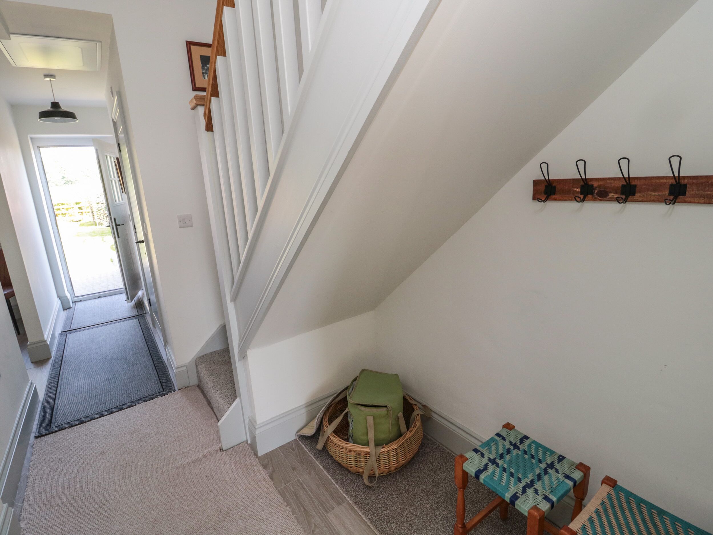 A staircase with a white railing, a door leading to a bright outdoor area, a wicker basket with a green cushion, and a woven chair with a blue and white cushion.