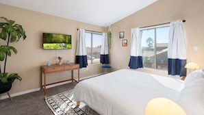 A bedroom with a bed, a TV mounted on the wall, a desk, and a window with curtains.