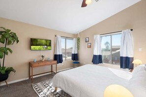 Ein Schlafzimmer mit einem Bett, einem an der Wand montierten Fernseher, einem Schreibtisch und einem Fenster mit Vorhängen.
