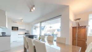 A modern kitchen with white cabinets and a dining area with a glass table and white chairs. - Lilly with parking by Interhome (Bredene)