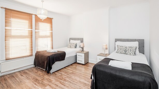 Ein modernes Schlafzimmer mit zwei Betten, Holzboden, weißen Wänden und einem großen Fenster mit Jalousien.