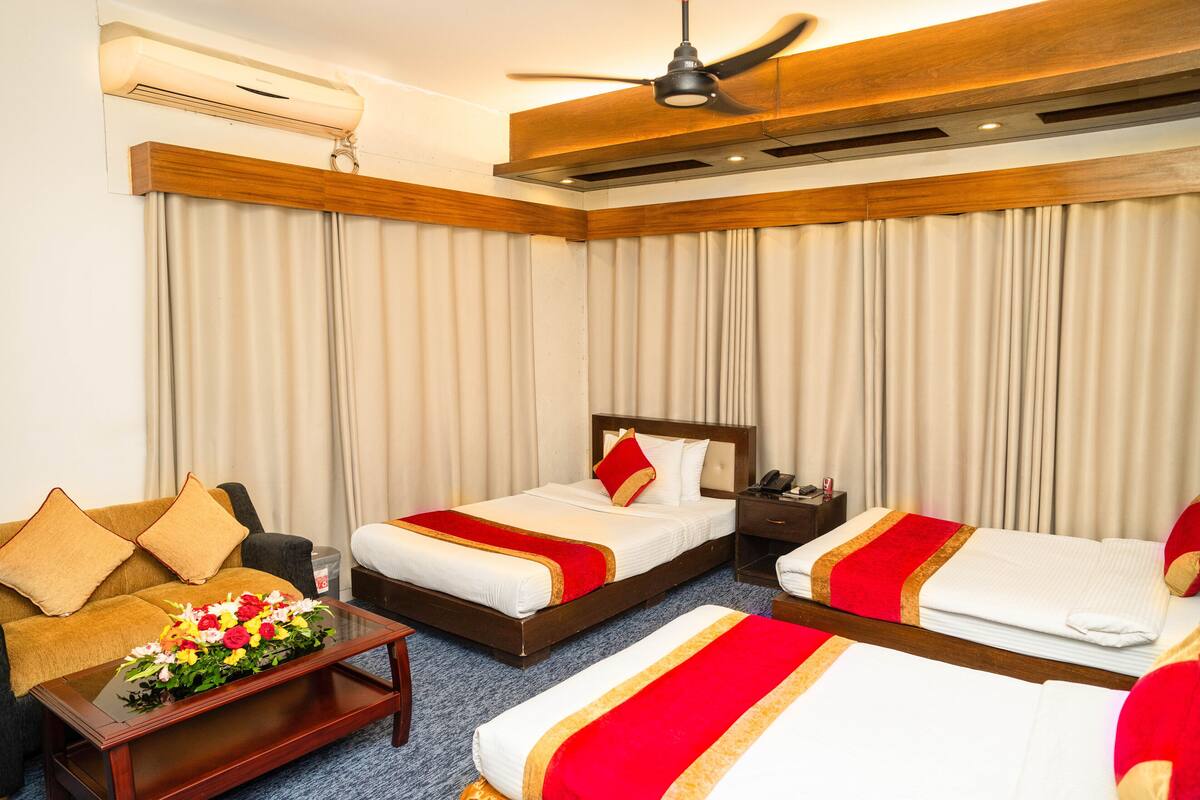 A hotel room with two beds, a sofa, a coffee table with flowers, an air conditioner, and a ceiling fan.