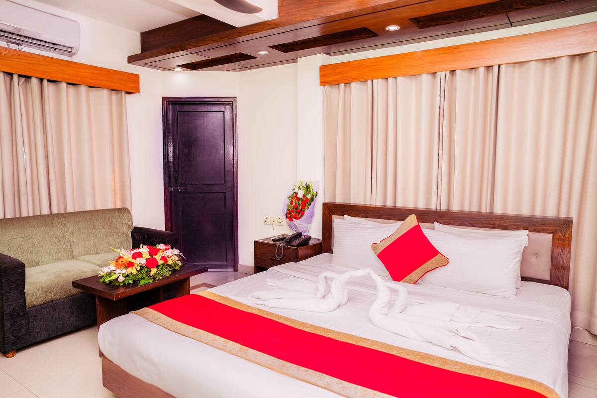 A hotel room with a large bed, a sofa, a nightstand with a vase of flowers, and a wooden headboard.