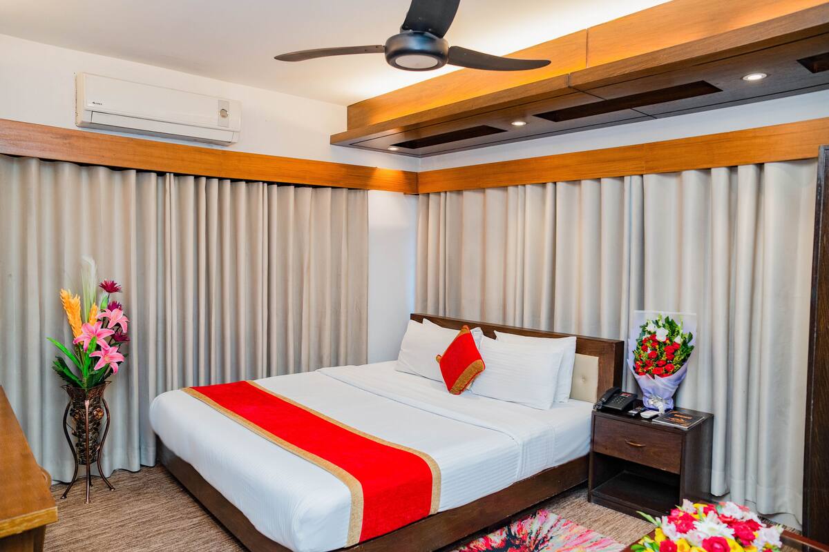 A hotel room with a large bed, wooden headboard, a ceiling fan, an air conditioning unit, a bedside table with a phone and a vase of flowers, and a decorative plant.