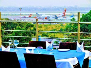 A dining area with tables set for two, overlooking an airport with multiple airplanes visible in the background. - Hotel Air Inn (Dhaka)