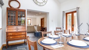 A dining area with a round wooden table set for six, surrounded by wooden chairs. A large wooden cabinet with glass doors is visible to the left. - Cwmwr (Oswestry)