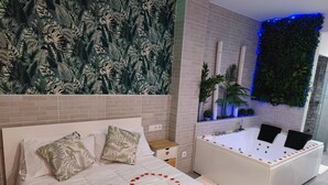 Une chambre moderne avec un grand lit, une décoration en forme de cœur faite de pétales de roses, une baignoire blanche et un mur vert orné de plantes.