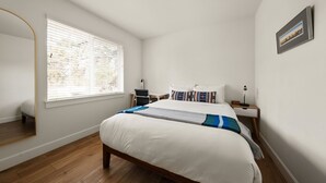 A bedroom with a bed, a bedside table, a chair, a window with blinds, a mirror, and a clock.
