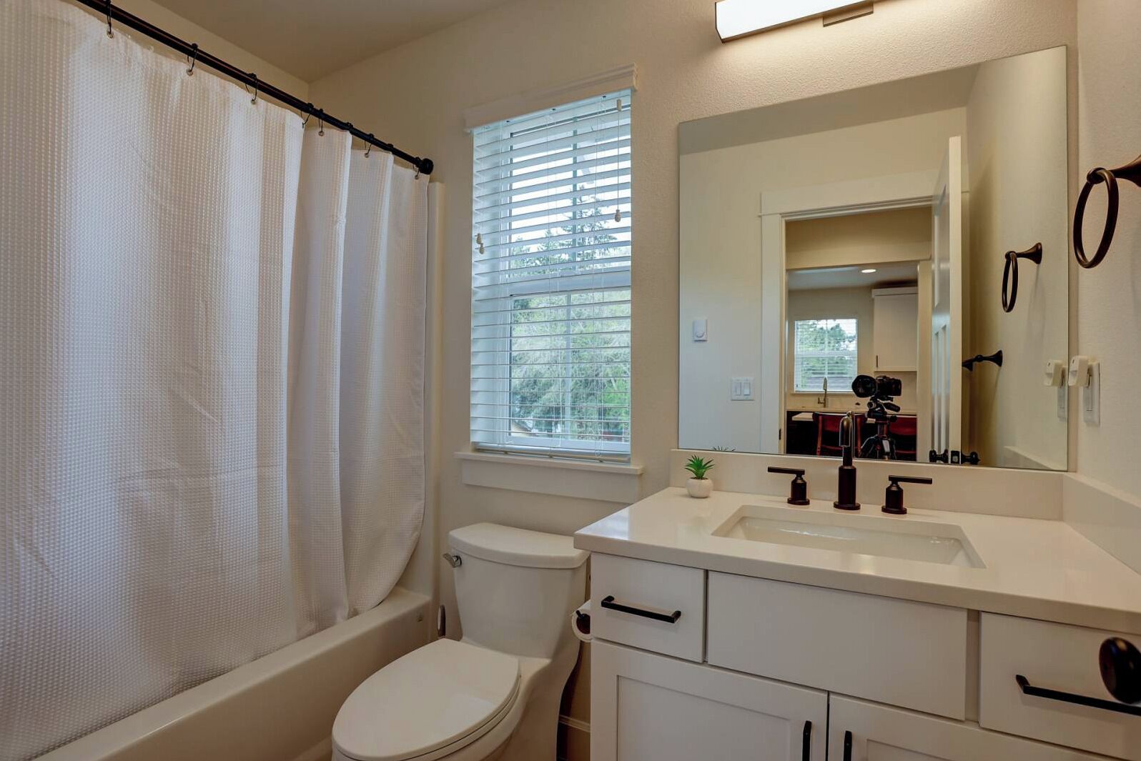 A bathroom with a toilet, a shower curtain, a window with blinds, a sink with a mirror, and cabinets.