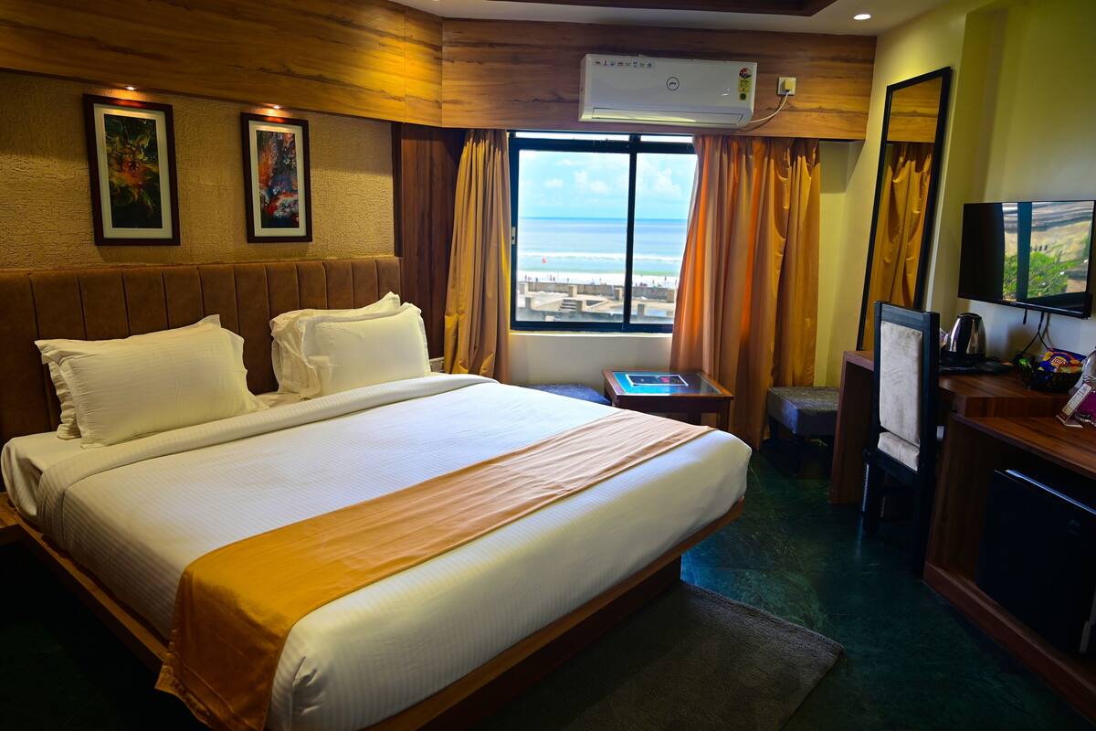 A hotel room with a large bed, a TV, a desk, and a window with a view of the beach.