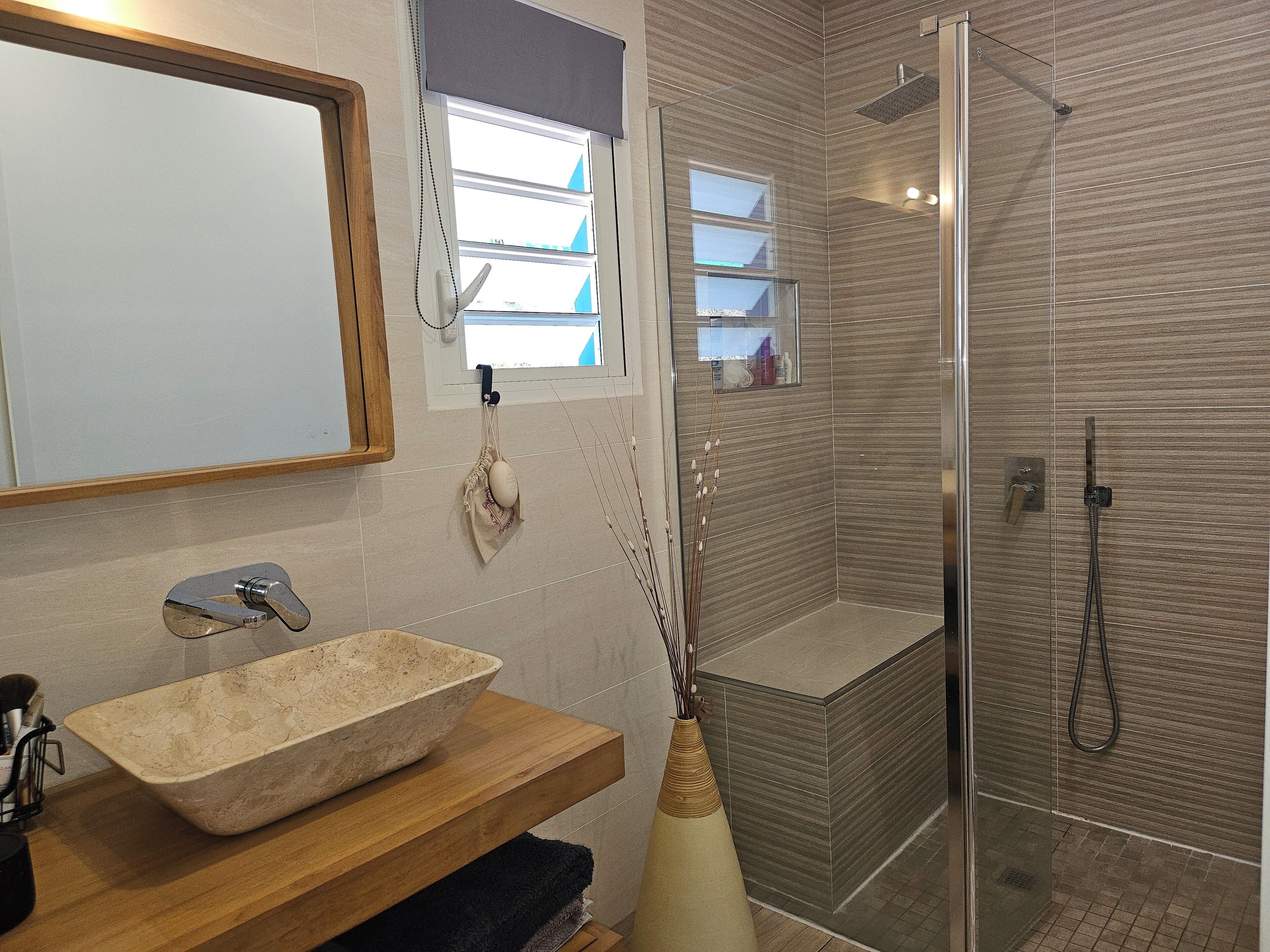 A bathroom with a stone sink, a mirror, a towel rack, a shower with a glass door, and a window with blinds.