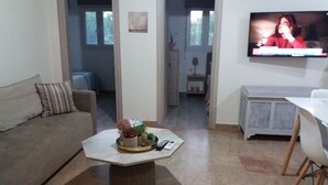 Una sala de estar con un sofá, una mesa de centro con adornos, un televisor en la pared y una zona de comedor con mesa y sillas.