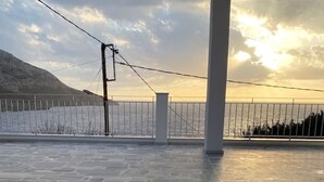 Een ruim terras buiten met een stenen vloer, een metalen hek en uitzicht op zee en bergen.