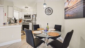 Una cocina moderna con armarios blancos, un refrigerador de acero inoxidable y un microondas. Hay un área de comedor con una mesa redonda y sillas negras, y un reloj de pared sobre la mesa.