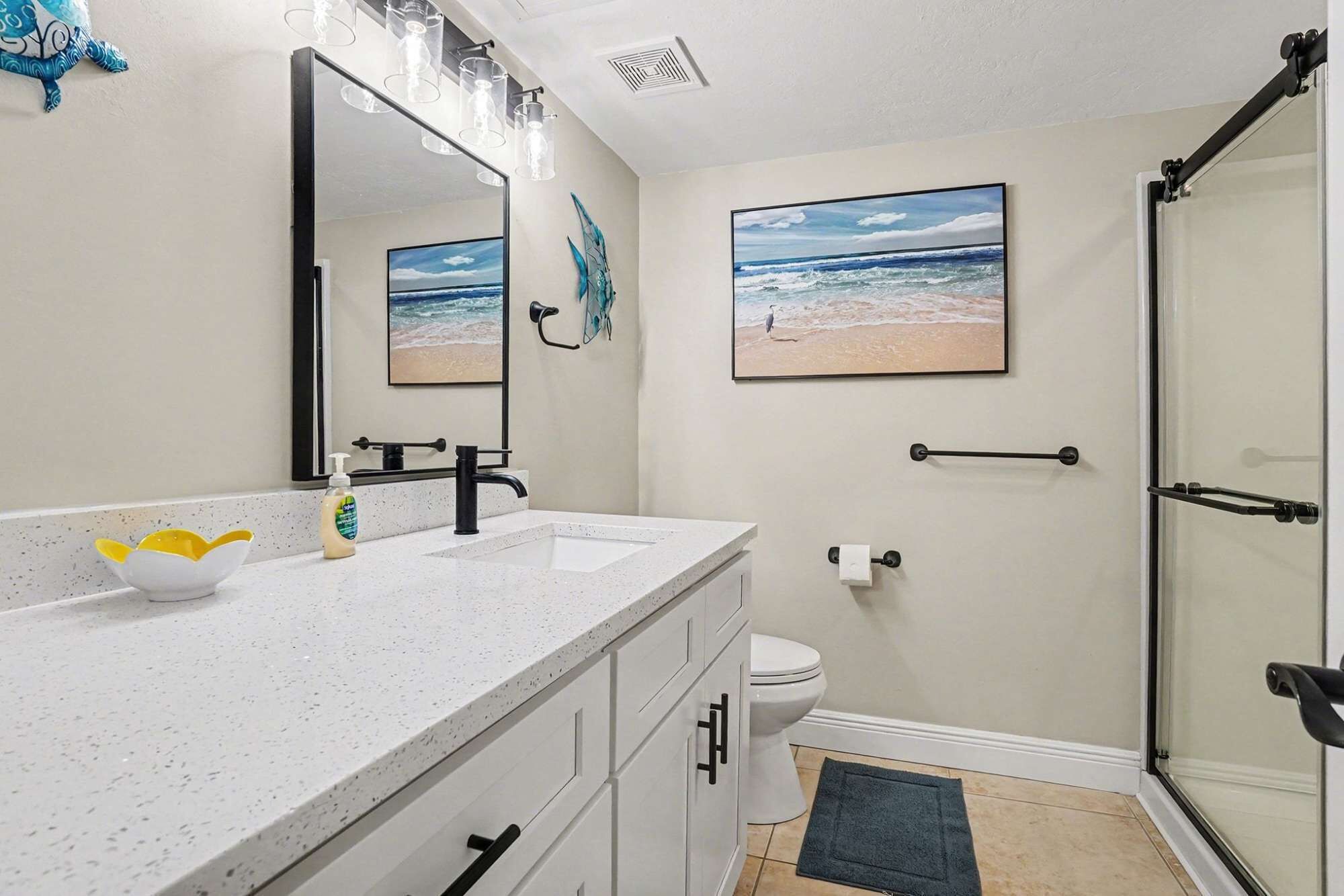 A bathroom with a white countertop, a large mirror, a shower with a glass door, and two framed pictures of beaches.