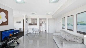A modern living room with a flat-screen TV, a sofa, a dining area, and a kitchen in the background.