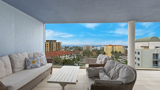A balcony with a sofa, a table, and a view of buildings and the sea.