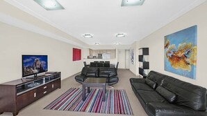 A modern living room with a flat-screen TV, leather sofas, a dining area, and a kitchen in the background.