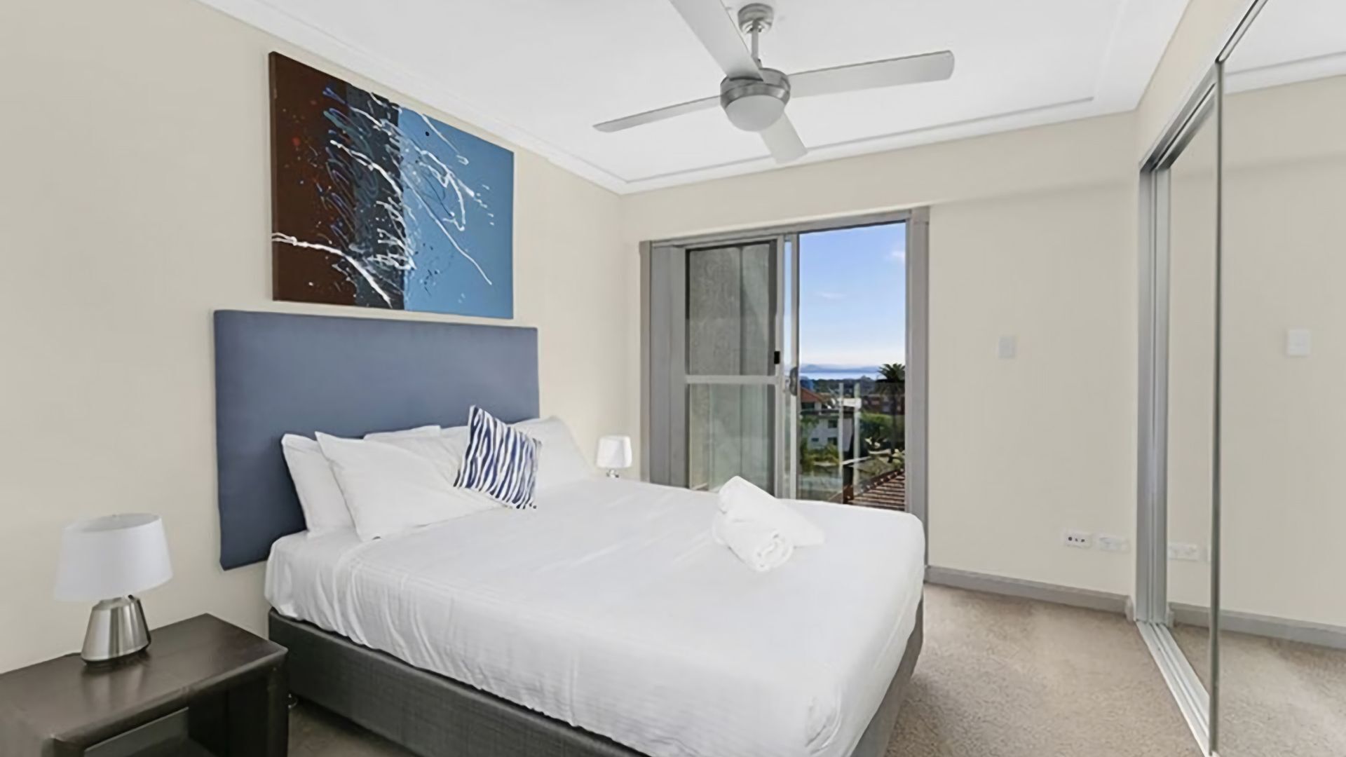 A bedroom with a bed, a ceiling fan, a nightstand, a lamp, a large painting, and a sliding glass door leading to a balcony.