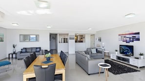 A modern living room with a dining area, grey sofas, a wooden dining table, and a TV on a white stand.