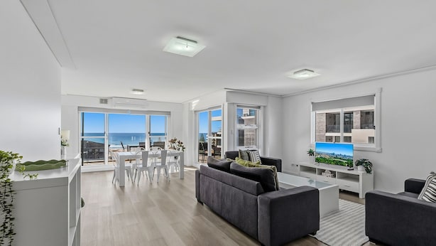 A modern living room with a grey sofa, a dining area with a table and chairs, and large windows offering a view of the sea. - Seaside Escape with Pool & Ocean Views (The Entrance)