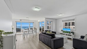 Un salon moderne avec un canapé gris, un coin repas comprenant une table et des chaises, et de grandes fenêtres offrant une vue sur la mer.